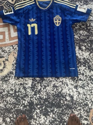 Blå Adidas sporttröja med mönster - Säljer en blå sporttröja från Adidas med unikt vågigt mönster i olika blå nyanser och gula detaljer vid halsen. Tillverkad i lätt och ventilerande material, perfekt för träning eller sportaktiviteter. Adidas-logga syns på insidan av kragen.