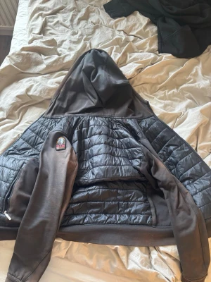 Svart vindjacka från Parajumpers - Snygg svart vindjacka från Parajumpers med quiltad kropp och släta ärmar. Jackan har huva, dragkedja och en patch på ärmen. Materialet är en mix av glansigt och matt tyg, vilket ger en modern look. Perfekt för dig som vill ha en stilren och funktionell jacka.