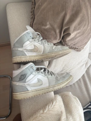 Nike Air Jordan 1 vita/grå - Säljer ett par Nike Air Jordan 1 sneakers i vitt och ljusgrått. Skorna har högt skaft, klassisk Jordan-logga på sidan och tungan, samt perforerad tåbox. Materialet är en mix av läder och textil. Snygga och tidlösa sneakers för dig som gillar streetwear. Storlek 46. Lite slitna bak i hälen men går att lösa och fixa med häl lappar. Säljer för 300kr