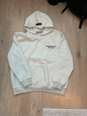 Grå Essentials Fear of God hoodie - Säljer en ljusgrå hoodie från Essentials Fear of God med svart logga tryckt på bröstet. Hoodien har en klassisk huva och ribbade muddar vid ärmar och nederkant. Perfekt för dig som gillar streetwear och enkel stil.