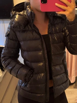 Moncler Bady hooded short down jacket - Snygg svart pufferjacka från Moncler med glansig finish, huva och dragkedja framtill. Moncler jacka i modellen Bady. Storlek 1 vilket motsvarar en xs. Finns QR-kod inuti som verifierar äkthet. Har även kvittot från farfetch där den är köpt. Mycket bra skick, inga tecken på användning. Nypris: 11 280kr