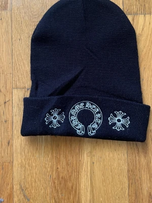 Svart mössa från Chrome Hearts - Svart ribbstickad mössa från Chrome Hearts med vit brodyr framtill. Mössan har uppvikt kant och detaljer med Chrome Hearts-logga och två kors. Perfekt accessoar för en edgy streetstyle-look.