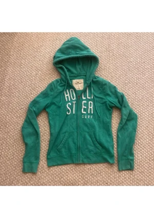 Grön zip hoodie från Hollister - Snygg grön hoodie från Hollister med dragkedja framtill och vit text på bröstet. Hoodien har två fickor, justerbar huva med snören och är gjord i mjukt bomullsmaterial. Perfekt för en avslappnad och chill stil.