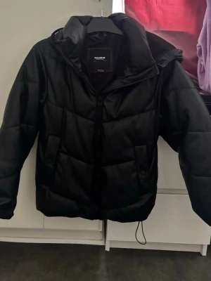 Svart pufferjacka med huva - Snygg svart pufferjacka från Pull&Bear med dragkedja framtill och stor huva. Jackan har en quiltad design och är vadderad för extra värme. Perfekt för kalla vinterdagar och har en modern, boxig passform. Använd i ett mycket bra skick storlek xs fast passar de som även har S.