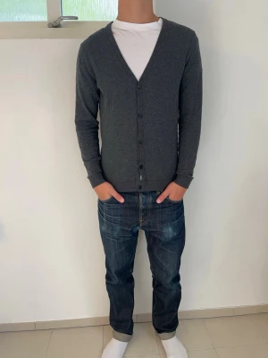 Mörkgrå cardigan från Selected Homme - Stilren mörkgrå cardigan från Selected Homme med v-ringning och knäppning framtill. Tröjan har långa ärmar och ribbade muddar vid ärmslut och nederkant. Perfekt att bära över en t-shirt för en avslappnad men snygg look. Storlek S men passar även till M. Hör av dig vid frågor!🙌