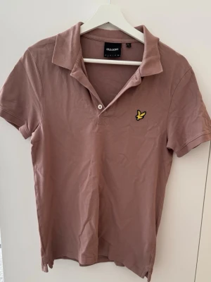 Rosa pikétröja från Lyle & Scott - Klassisk kortärmad pikétröja från Lyle & Scott i en stilren dusty pink / brunrosa nyans. Tröjan har krage, knäppning framtill och den ikoniska gula örn-logotypen broderad på bröstet. Perfekt för en fräsch och uppklädd vardagsstil.