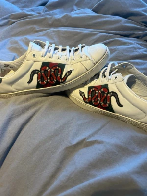 Äkta Vita sneakers med orm brodyr -  Äkta Snygga vita sneakers i läder med broderad orm i rött, svart och vitt på sidan samt grönt och rött band bakom ormen. Klassisk låg modell med vita skosnören och gummisula. Perfekta för dig som vill sticka ut med cool detalj.