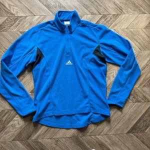 Blå långärmad träningsfleece Adidas - Blå långärmad träningströja från Adidas med halv dragkedja framtill och svarta detaljer vid sidorna. Tillverkad i ett mjukt funktionsmaterial som passar perfekt för löpning eller annan träning. Adidas-logga på bröstet och hög krage för extra komfort.