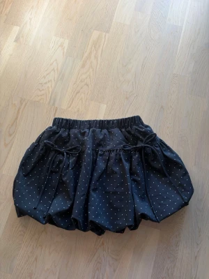 Svart kortkjol med prickigt mönster - Söt svart kortkjol med små vita prickar och voluminös ballongform. Kjolen har resår i midjan och dekorativa knytband framtill. Köpte denna i en liten butik i SydKorea för 1 månad sedan så den ör aldrig använd endast testad🥰