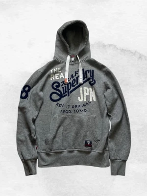 Grå hoodie från Superdry med tryck - Snygg grå hoodie från Superdry med stort blått och vitt tryck på bröstet och ärmen. Klassisk känguruficka framtill, vita dragsnören i huvan och ribbade muddar. Perfekt för en avslappnad streetstyle-look.