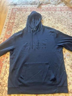 Mörkblå hoodie från Hugo Boss - Snygg mörkblå hoodie från Hugo Boss med broderad logga på bröstet. Klassisk känguruficka framtill, justerbar huva med dragsko och ribbade muddar. Perfekt för en avslappnad och stilren look.