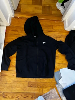 Svart zip hoodie från Nike - Snygg svart hoodie med dragkedja från Nike. Klassisk design med vit Nike-logga på bröstet, huva och två stora fickor framtill. Perfekt för en avslappnad och sportig stil. Tillverkad i mjukt material för extra komfort.