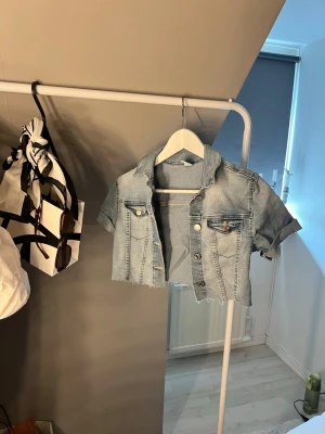 Ljusblå croppad jeansjacka - Trendig ljusblå croppad jeansjacka med korta ärmar och rå kant nertill. Klassisk krage och två bröstfickor med knappar framtill. Perfekt för en cool och avslappnad stil.