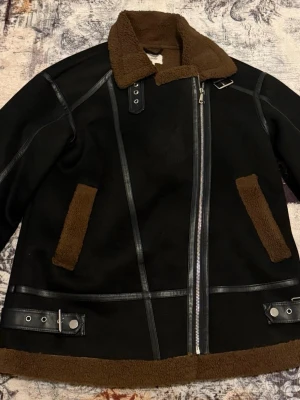 Svart teddyfodrad pälsjacka / Orginal pris 1200kr - Säljer en svart pälsjacka med brunt teddyfoder och detaljer. Jackan har bred krage, dragkedja framtill, bältesdetaljer vid krage och nederkant samt stora fickor med brunt foder. Perfekt för kalla vinterdagar och riktigt snygg till streetwear.