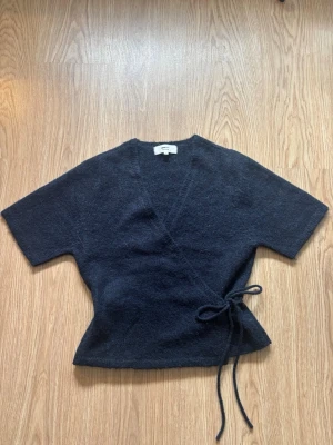 Mörkblå omlottopp med knytband - Snygg mörkblå omlottopp med korta ärmar och knytband i midjan. Tröjan har en mjuk, lite luddig textur och en feminin siluett. Perfekt att styla med jeans eller kjol för en trendig look.