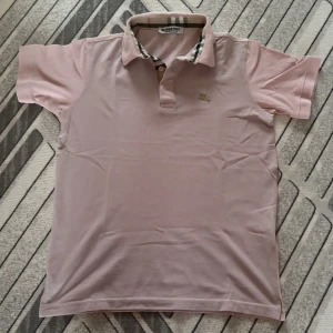 Rosa burberry pike - Snygg ljusrosa pikétröja med korta ärmar och klassisk krage. Kragen har ett rutigt foder som ger en extra detalj. Tröjan har knappar framtill och en liten broderad logga på bröstet. Perfekt till jeans eller shorts.