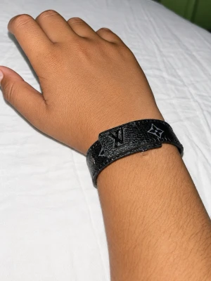 Svart Lv läderarmband med monogram - Snyggt svart armband i läder med monogrammönster och silverfärgad detalj. Armbandet har en stilren design och justerbart spänne. Perfekt accessoar för dig som gillar en clean och exklusiv look.