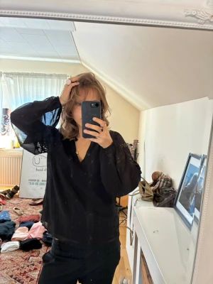 Svart blus med spetsdetaljer - Snygg svart blus med v-ringning och transparenta, vida långärmar. Blusen har fina spetsdetaljer på ärmarna som ger en trendig touch. Tillverkad i ett lätt och luftigt material som faller snyggt.