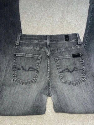 Grå bootcut jeans med broderade fickor - Säljer ett par grå jeans med bootcut och snygga broderade detaljer på bakfickorna. Jeansen har klassisk femficksdesign och bälteshällor. Perfekta för en avslappnad och stilren look. Storlek XXS men passar även XS. Innerbenslängd: 79cm. Slitna längst ned (se bild) 