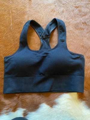 Svart sport-bh med brottarrygg - En svart sport-bh i ribbat material med bred resår under bysten och brottarrygg. Bh:n har en enkel, stilren design utan synliga märken eller dekorationer. Perfekt för träning eller en aktiv vardag. Använd en gång