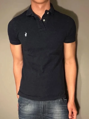 Svart pikétröja från Ralph Lauren - Klassisk svart pikétröja från Ralph Lauren med vit broderad logga på bröstet. Tröjan har korta ärmar, krage och knappar vid halsen. Tillverkad i mjuk bomull som ger en bekväm och snygg passform.