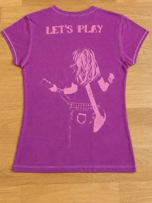 Lila Hard Rock Cafe t-shirt - Lila t-shirt från Hard Rock Cafe Barcelona med tryck fram och bak. Framsidan har logga, text och biljettmotiv, medan baksidan har ett coolt motiv av en person med gitarr och texten 'LET'S PLAY'. Figurnära passform och korta ärmar. Perfekt för musikälskare! Jag skulle säga att den är krympt lite.