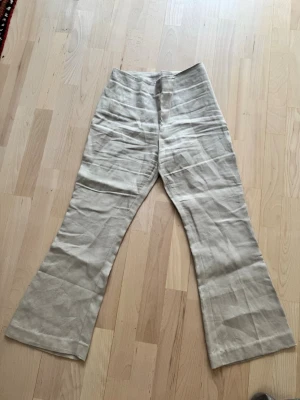 Beiga vida linnebyxor med dragkedja - Snygga beiga linnebyxor med hög midja och vida ben. Byxorna har en diskret dragkedja i sidan för en clean look. Perfekta för en chill och stilren outfit under varma dagar. Materialet är svalt och luftigt linne.