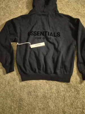 Svart Essentials Fear of God hoodie - Säljer en svart hoodie från Essentials Fear of God med stor logga på ryggen och mindre logga på bröstet. Hoodien har huva och en klassisk magficka framtill. Tillverkad i mjukt material som passar perfekt för en avslappnad streetwear-look. Hoodien är helt ny och aldrig använd. STORLEKEN ÄR XXS OCH PASSAR FOLK SOM ÄR CA 165CM och längre. 