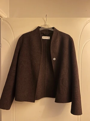 Brun jacka i ullmix från Wera - Midjekort jacka med rena, avskalade linjer, tillverkad i varm och härlig ullmix. NIKE har rak passform, kraglös hals med V-ringning och raka ärmar. Jackan stängs med en dold knapp upptill och på framsidan sitter två dolda fickor. Köpt på Åhléns för 1999kr🧡