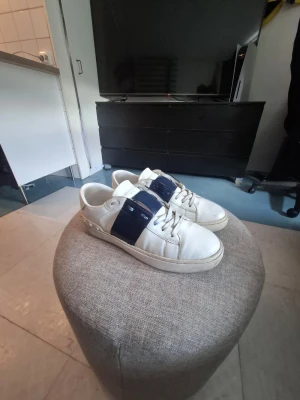 Valentino - Snygga vita sneakers med breda mörkblå band över vristen och vita snören. Skorna har en klassisk låg profil och rundad tå. Tillverkade i slätt läder med gummisula för skön komfort. Perfekta för en clean och stilren look.
