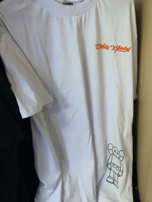 Off-White x KAWS vit t-shirt - Vit t-shirt från Off-White x KAWS med tryck i orange och svart framtill. Framsidan har en liten Off-White x KAWS-logga på bröstet och en stor KAWS-figur med detaljer i orange och svart på baksidan. Tillverkad i mjuk bomull och har en oversized passform.
