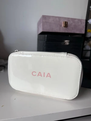 Vit necessär från CAIA - Snygg vit necessär från CAIA i glansigt material med dragkedja runtom. Perfekt för smink eller småprylar. Enkel och stilren design med rosa CAIA-logga på framsidan. Praktisk storlek att ha med sig i väskan.