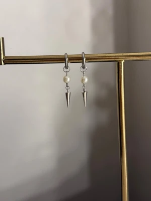 Silvriga örhängen med pärlor och spikes - Säljer ett par stilrena örhängen i silverfärg med små vita pärlor och spetsiga spikes som hänger ned. Örhängena har en modern och edgy look med en mix av klassiska och tuffa detaljer. Perfekta för dig som gillar att sticka ut med dina accessoarer.