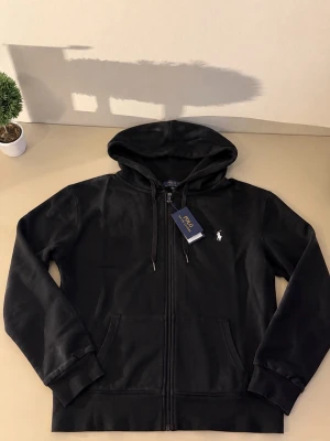 Svart hoodie från Polo Ralph Lauren - Snygg svart hoodie från Polo Ralph Lauren med dragkedja framtill och klassisk huva. Ikoniska vita loggan broderad på bröstet. Tillverkad i mjukt bomullsmaterial och har två fickor framtill. Perfekt för en avslappnad och stilren look.