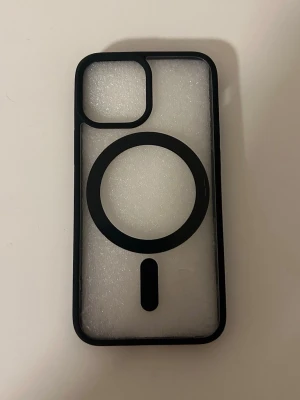 Svart MagSafe-kompatibelt mobilskal för iphone 13 mini - Snyggt svart mobilskal med MagSafe-kompatibilitet. Skalet har en genomskinlig baksida och en markerad cirkel för MagSafe-funktion. Tillverkat i tålig plast med exakta utskärningar för kamera och knappar. Perfekt för dig som vill ha ett stilrent och funktionellt skydd till din mobil.