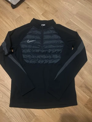 Svart träningsjacka från Nike - Svart långärmad träningsjacka från Nike med halv dragkedja och vadderad frontpanel. Jackan har en diskret Nike-logga på bröstet och gråa detaljer längs ärmarna. Perfekt för löpning eller träning utomhus under kyligare dagar.
