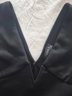 Svart kort klänning från Club L - Säljer en svart, figurnära kort klänning från Club L med djup v-ringning. Klänningen har en enkel och stilren design utan ärmar och är tillverkad i ett mjukt, stretchigt material som sitter snyggt på kroppen.