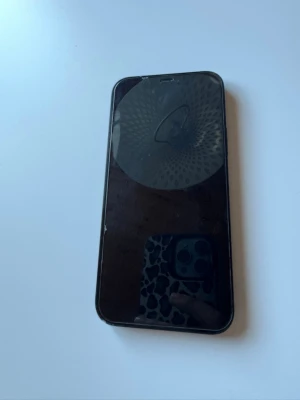 IPhone 12 - Säljer en svart iPhone med stilren design och rundade kanter. Telefonen har två kameror på baksidan och den klassiska Apple-loggan. Baksidan är i glas och ramen är i metall. Perfekt för dig som vill ha en snygg och modern smartphone.