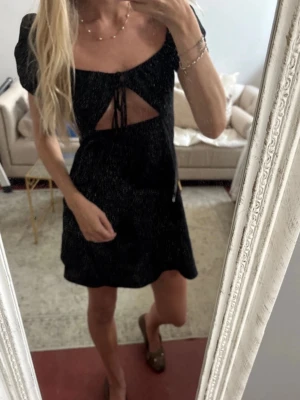 Svart kort klänning med snörning - Svart kort klänning med puffärm och snygg cut-out framtill som knyts med snörning. Klänningen har en feminin siluett och är perfekt för dig som vill ha en trendig och stilren look. Materialet ser ut att vara mjukt och följsamt.