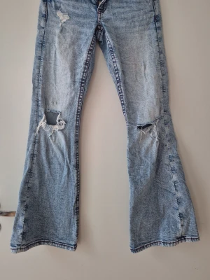 Ljusblå slitna bootcut jeans Gina Tricot - Ljusblå bootcut jeans från Gina Tricot med slitningar och hål på båda knäna. Jeansen har röd kontrastsöm och klassisk femficksmodell. Perfekta för en avslappnad och trendig look.  På sista bilden finns det en svag fläck som jag inte försökt att tvätta bort. Hund finns i hemmet. 