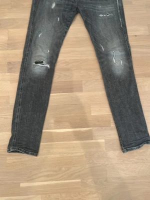 Grå slitna jeans med raka ben - Säljer ett par grå jeans med slitningar och hål på framsidan. Jeansen har raka ben och klassisk femficksdesign. Materialet är denim och färgen är tvättad grå med ljusa partier och distressed detaljer för en cool, avslappnad look. Modell Ambass slimfit. En lagning på vänstersidan men flyter in i övriga slitna detaljer. 