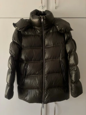 Mörkgrön pufferjacka med huva - Säljer en slutsåld mörkgrön pufferjacka från massimo dutti, med glansig finish och stor huva. Jackan har två dragkedjeförsedda fickor framtill och breda, vadderade paneler för extra värme. Perfekt för kalla vinterdagar och riktigt snygg till streetwear-stilen.