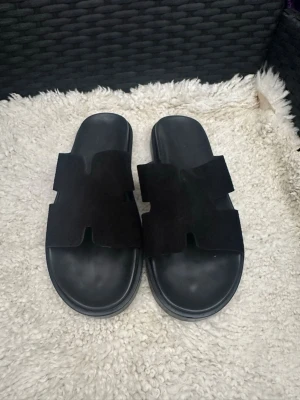 Svarta slip-in sandaler i mocka - Stilrena svarta slip-in sandaler med breda remmar i mocka och bekväm sula. Perfekta för varma dagar och enkel att matcha till olika outfits. Klassisk design med öppen tå och tidlös look.