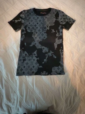 Svart och grå mönstrad t-shirt - Säljer en svart och grå t-shirt med korta ärmar och ett unikt mönster som blandar camouflage och loggor. T-shirten har en slim passform och är tillverkad i ett mjukt material som känns skönt mot huden. Perfekt för dig som vill sticka ut med en cool look.