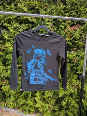 Långärmad tröja strl 122/128 lego ninjago - Svart tröja med lite blå skimrande tryck  Tyvärr fått ett litet jack på ena ärmslutet - se bild  100% bomull 