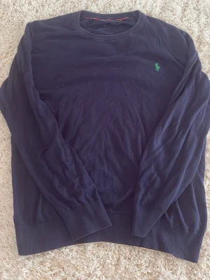 Mörkblå tröja från Ralph Lauren - Klassisk mörkblå tröja från Ralph Lauren med rund halsringning och den ikoniska gröna logotypen broderad på bröstet. Tröjan har långa ärmar och ribbade muddar vid ärmslut och nederkant. Perfekt för en stilren och enkel look. Står storlek L, men passar mer som M