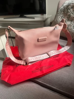 Rosa axelväska från Valentino - Snygg rosa axelväska från Valentino med bred axelrem där loggan är tryckt i vitt. Väskan har en stilren, rektangulär form och är tillverkad i ett slätt, veganskt lädermaterial. Dragkedja upptill och diskret logotyp framtill.