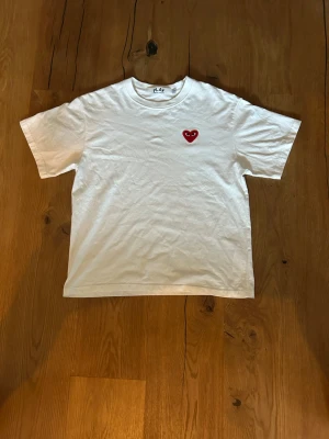 Vit t-shirt med rött hjärta - Vit t-shirt med ett litet rött hjärta broderat på vänster bröst. Klassisk passform och rund halsringning. Perfekt basic-plagg med en söt detalj framtill. Tillverkad i mjuk bomull för skön känsla.
