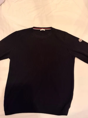 Svart stickad tröja från Moncler - Snygg svart stickad tröja från Moncler med rund halsringning och Moncler-logga på ena ärmen. Tröjan har en klassisk passform och är ribbstickad för extra struktur. Perfekt för dig som gillar stilrena och exklusiva plagg.