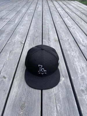 LA keps - Säljer en keps från New Era med LA-logga broderad framtill i vitt. Klassisk 59FIFTY-modell med platt skärm och ventilation på ovansidan. Insidan har märkets band och etiketter. Perfekt för dig som gillar streetwear och sportig stil. Storlek: 59,6 cm